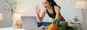 A woman in a sleeveless top works on her laptop, greeting someone while surrounded by fresh vegetables and fruits on the table.