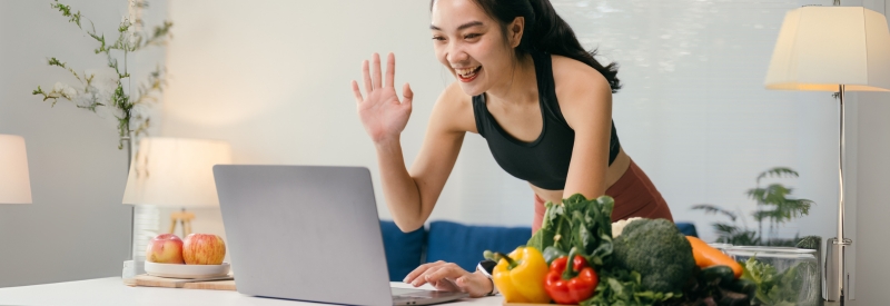 A woman in a sleeveless top works on her laptop, greeting someone while surrounded by fresh vegetables and fruits on the table.